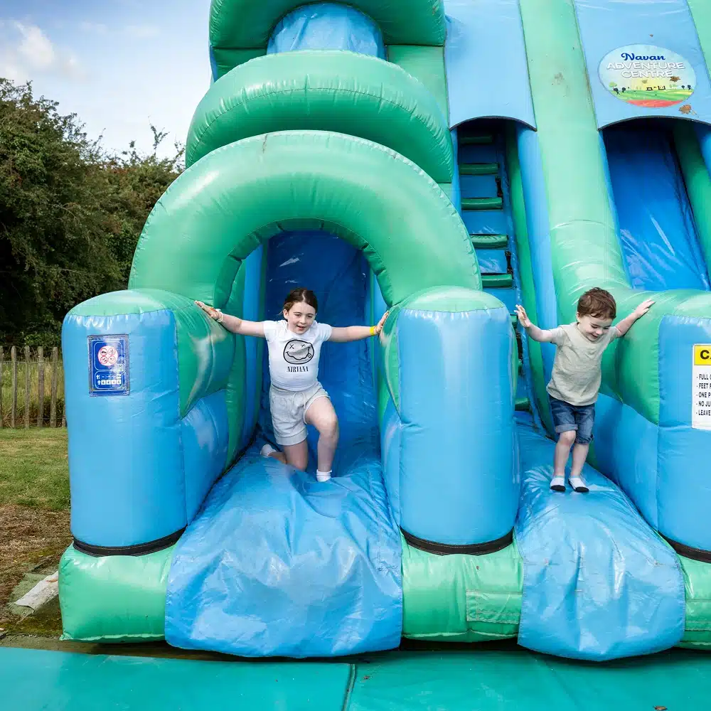 Two children happily exit an inflatable blue and green slide at NeuroFest Navan. The girl on the left kneels and smiles, while the boy on the right steps off the slide. Trees and grass are visible in the background.