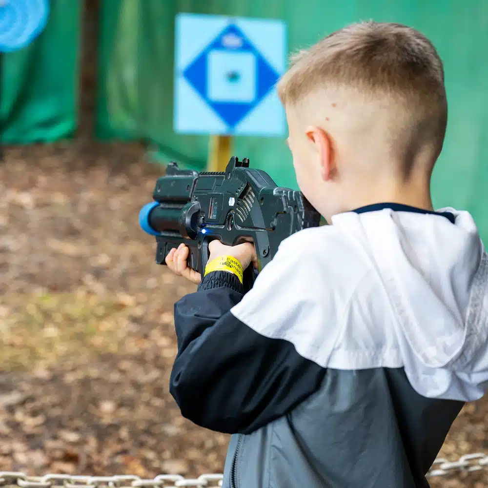 At NeuroFest Navan, a young boy in a grey and white jacket aims a toy gun at a blue and white target outdoors, with a blurred background of leaves and green panels.