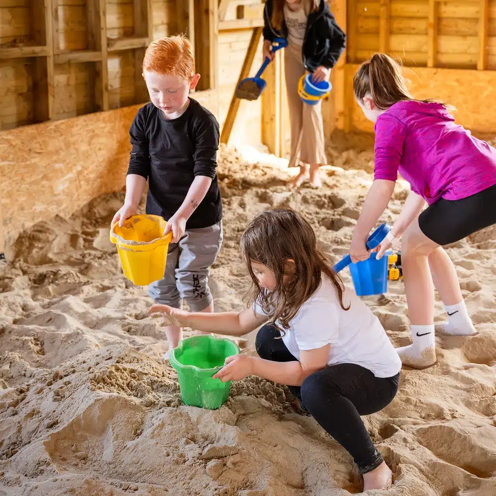 Four children play in an indoor sandpit at Navan Easter camp, scooping sand with colourful buckets. Two kids are standing, while two are crouched down, all focused on their activity as sunlight streams through the wooden walls.