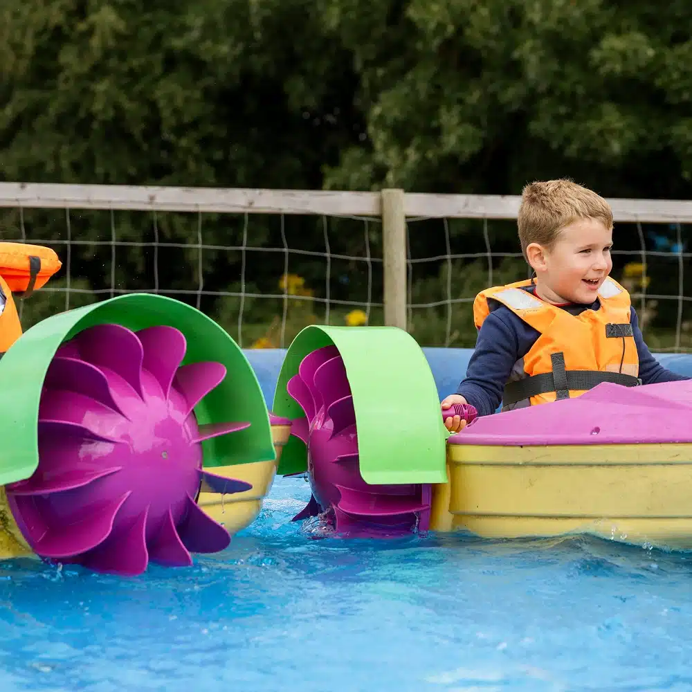 A young boy in an orange life jacket smiles whilst sitting in a colourful paddle boat on a pool at Navan Easter camp, with trees and a wire fence in the background.