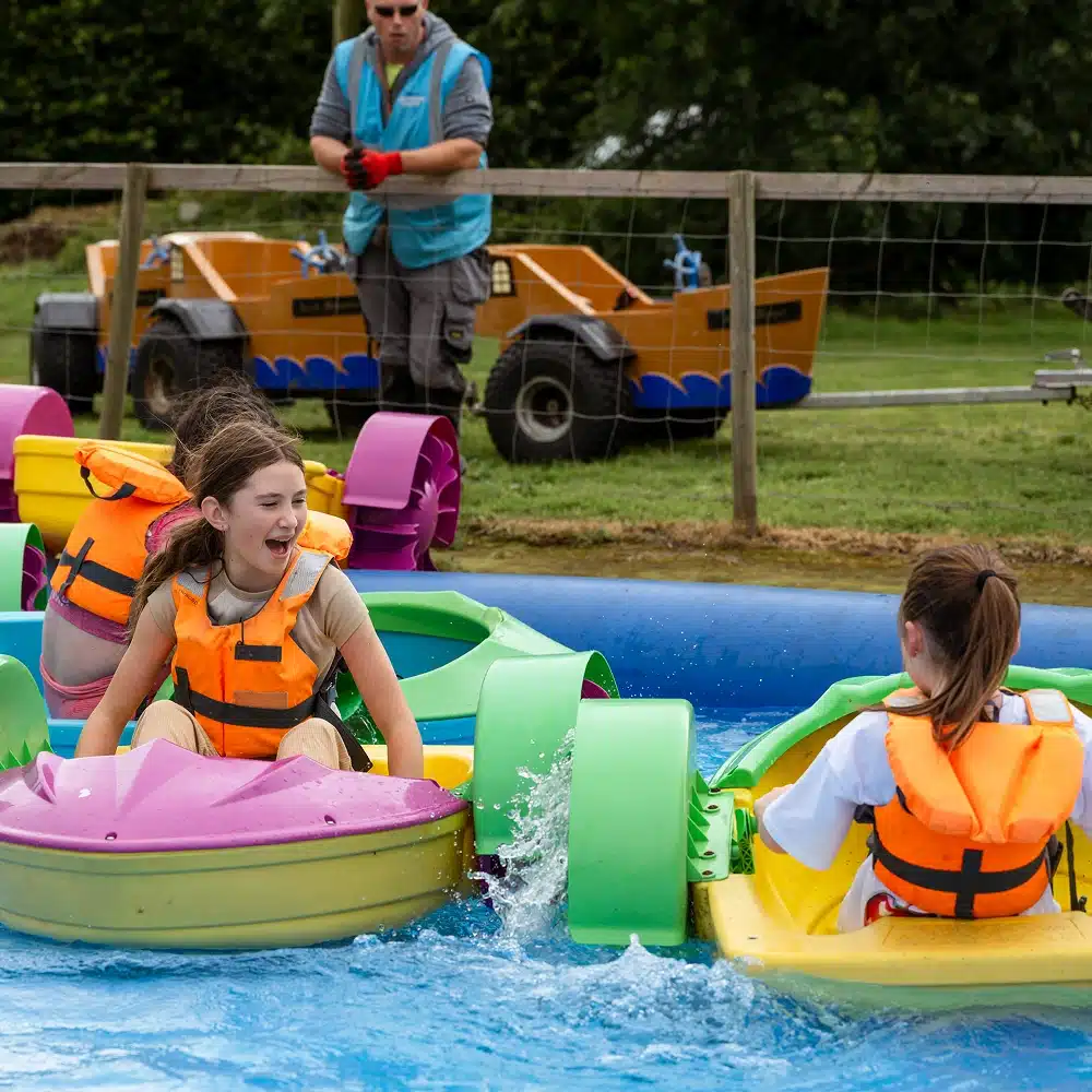 Two children wearing orange life jackets paddle small colourful boats in a shallow pool at Navan Easter camp. Another person in a blue vest stands nearby on the grass, with equipment and fencing visible in the background.