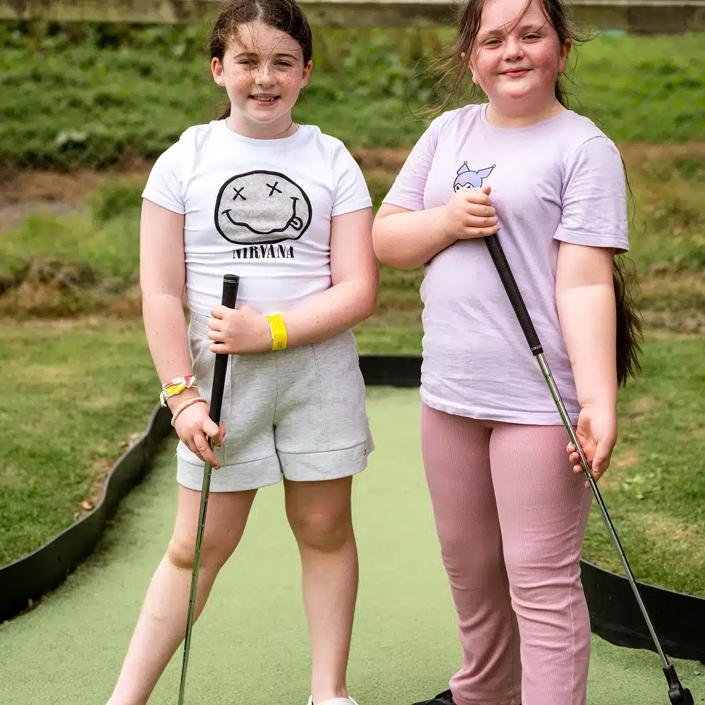 Two young girls at Navan Easter camp stand on a mini-golf course, holding golf clubs and smiling at the camera. One wears a Nirvana t-shirt and shorts, while the other wears a purple shirt and trousers. Grass and a fence are in the background.