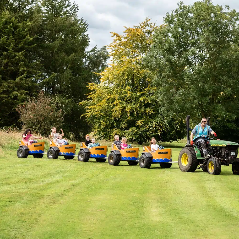 At NeuroFest Navan, a man drives a green tractor pulling yellow carts, each carrying a child riding through the grassy area surrounded by trees. The children appear to be enjoying the ride.