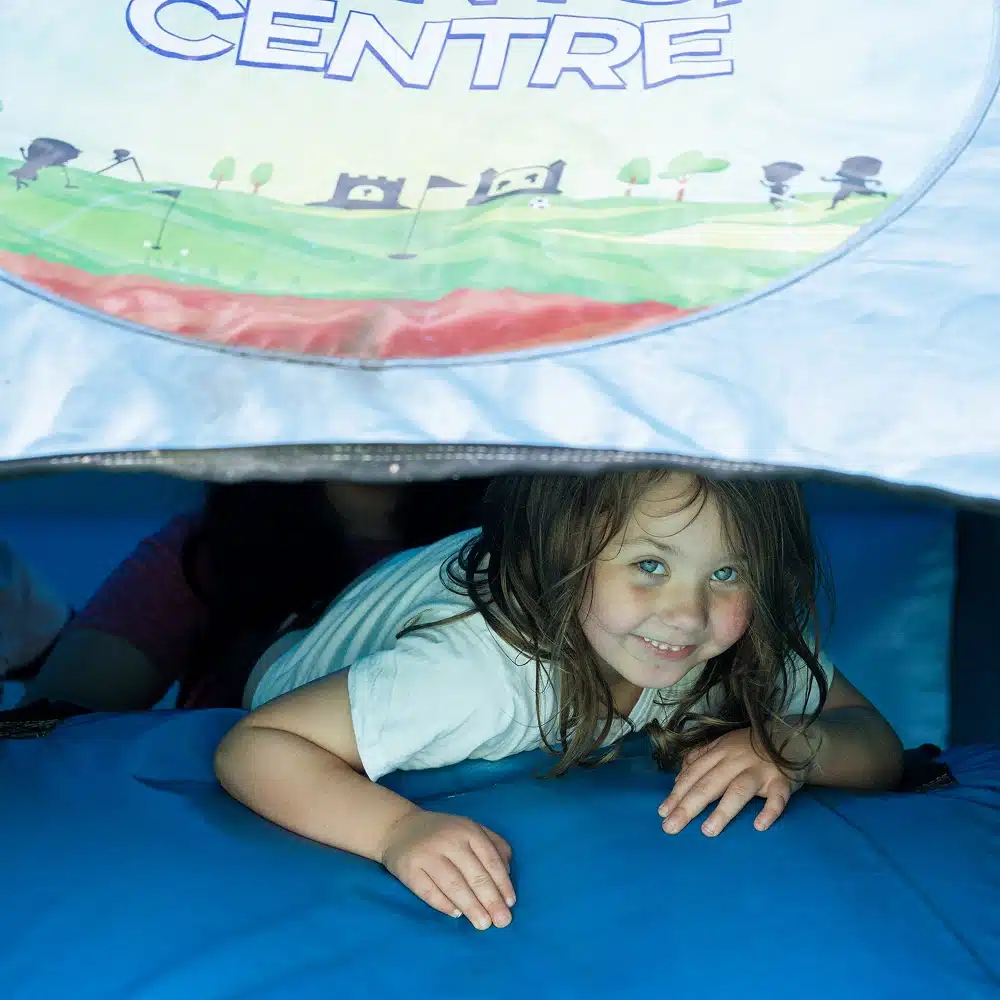 A young child with long hair and a white shirt smiles whilst crawling under a blue tent with colourful designs and the word "CENTRE" visible above at Navan Easter camp.
