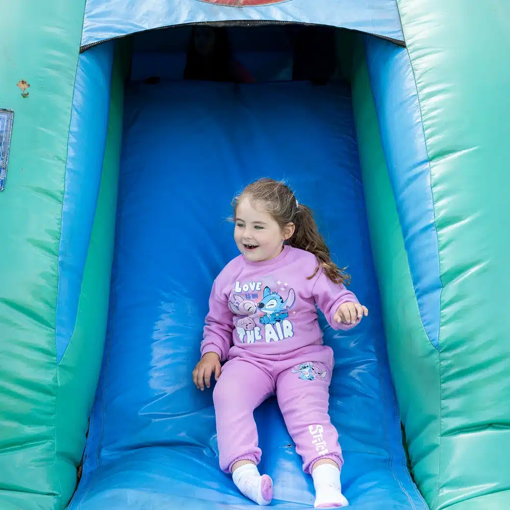 A young girl in a pink outfit slides down a blue and green inflatable slide at Navan Easter camp, smiling with her hair in a ponytail.