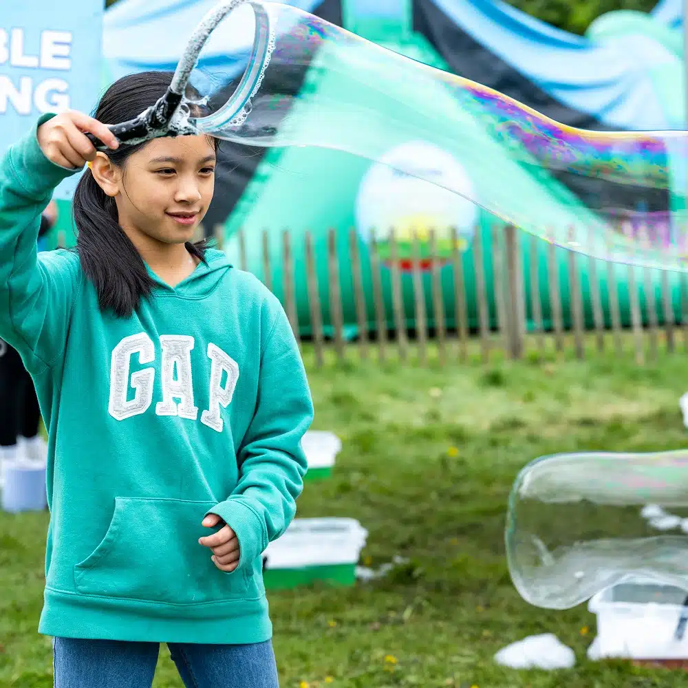 At Navan Easter camp, a girl in a green GAP hoodie makes a large soap bubble outdoors using a bubble wand. She stands on grass with a blurred fence and colourful play equipment in the background.