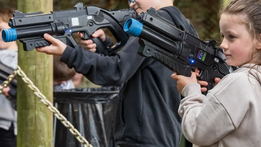Two children, a boy and a girl in casual clothes, aim large toy laser guns while enjoying an indoor laser tag game at Navan Easter camp. The background features ropes, wooden posts, and blurred greenery.