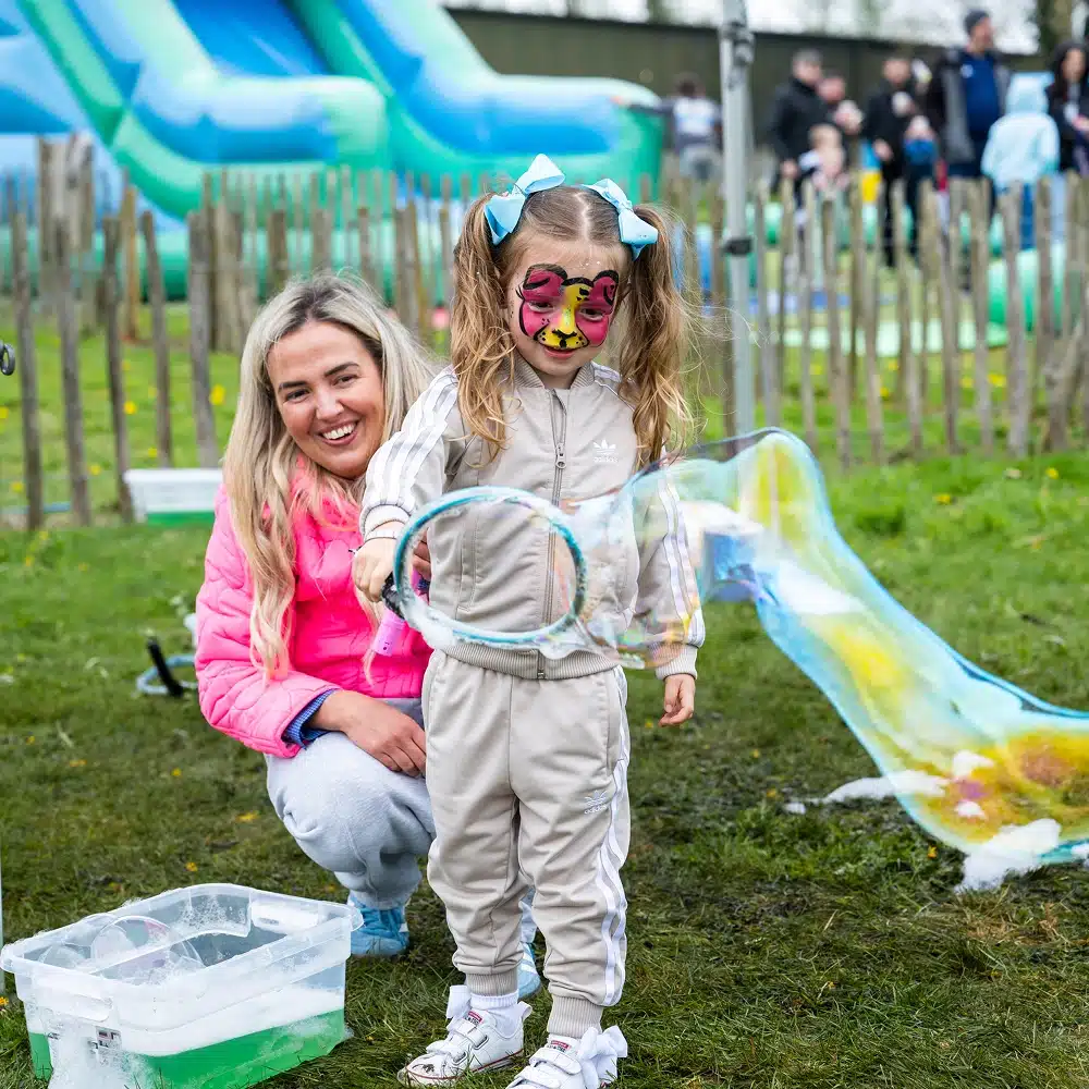 A young girl with face paint and blue bows blows large bubbles outdoors at the Navan Easter event, holding a bubble wand. An adult kneels nearby, smiling, whilst people and inflatable slides are visible in the background.