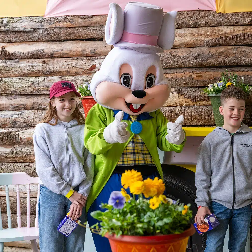Two smiling children stand beside a person in a white bunny costume with a green coat and top hat at the Navan Easter event, posing outdoors near colourful flowers and a wooden background.