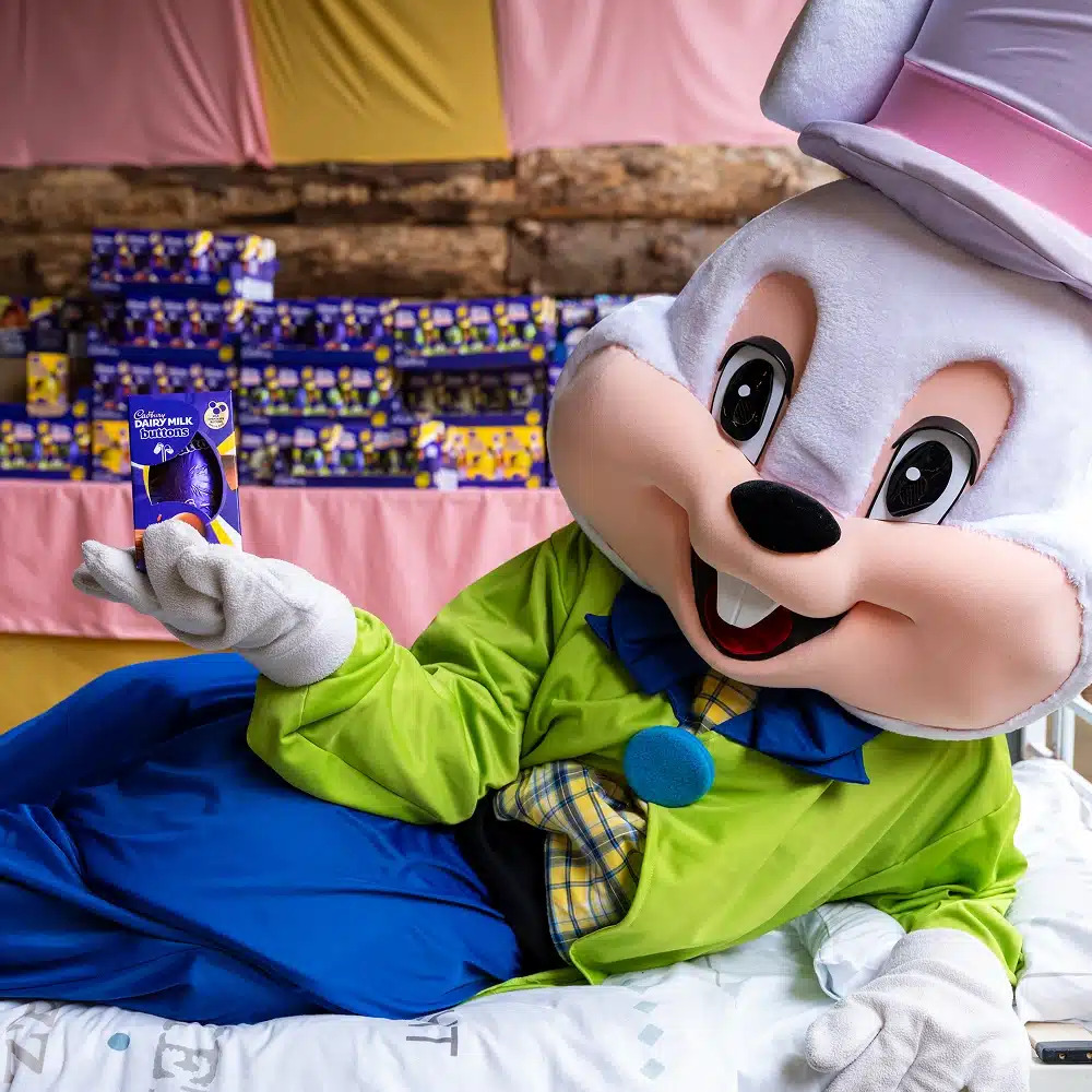 A person in a large Easter bunny costume, wearing a green jacket and pink hat, reclines on a bed whilst holding a Cadbury Dairy Milk chocolate egg at the Navan Easter event. Stacks of Cadbury Easter egg boxes are visible in the background.