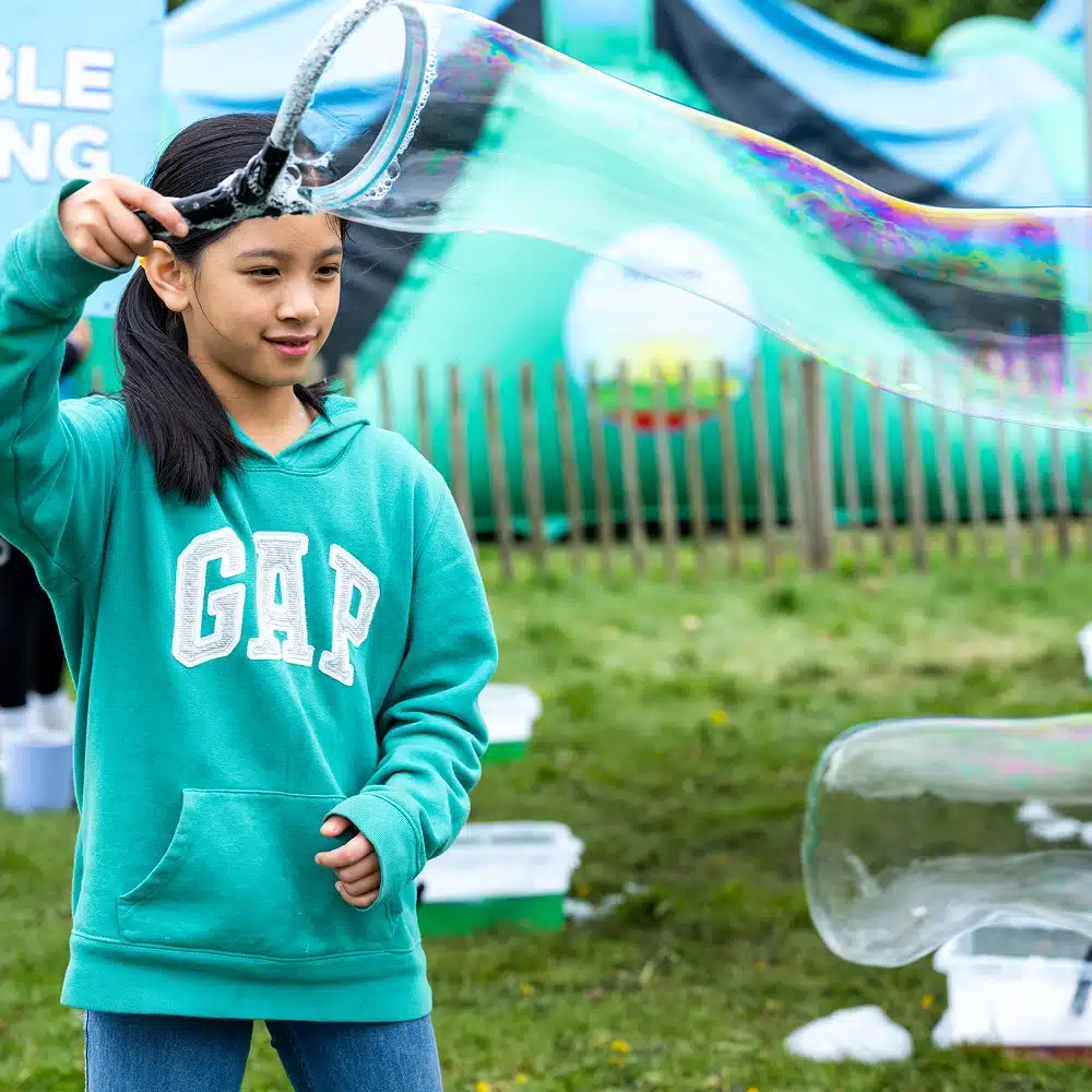 At the Navan Easter event, a young girl in a green GAP hoodie creates a giant bubble with a wand on the grass, surrounded by bubble-making supplies and a blurred inflatable attraction in the background.
