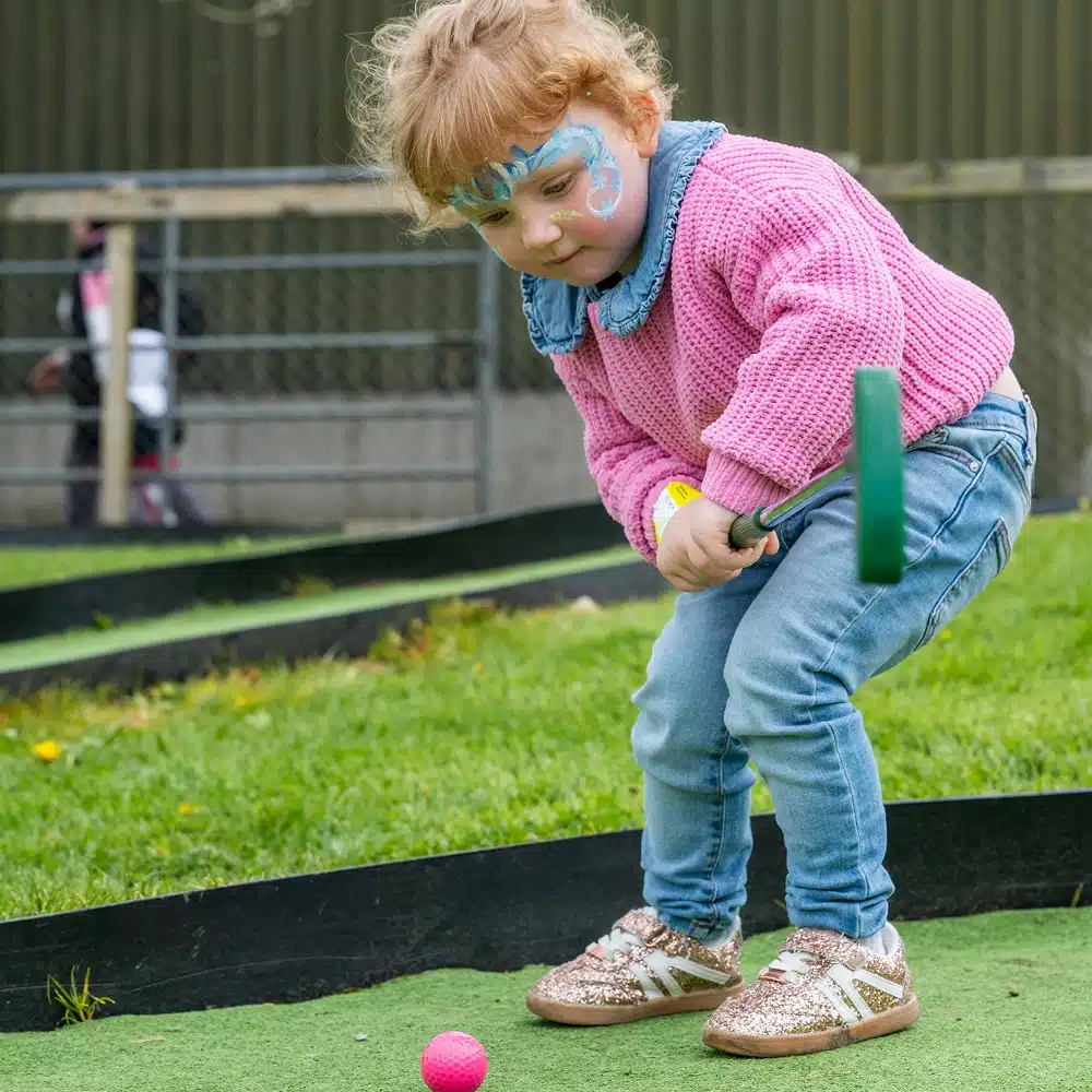 A young child with face paint, wearing a pink jumper and jeans, is playing crazy golf outside at the Navan Easter event. They are bending down, focusing on hitting a pink golf ball with a green club on the grassy course.