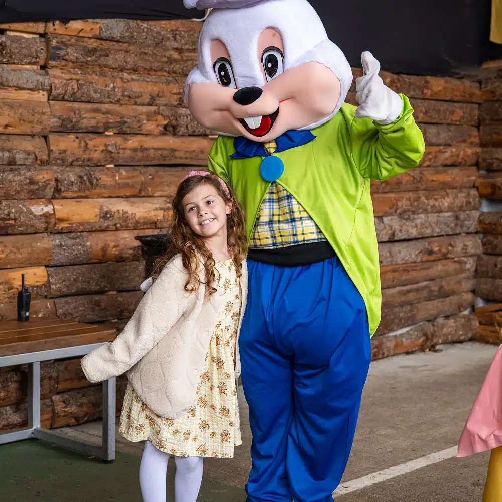 A smiling girl in a floral dress and cream jacket poses next to a person in a large white bunny costume with green and blue clothes at the Navan Easter event, standing in front of a wooden log wall.