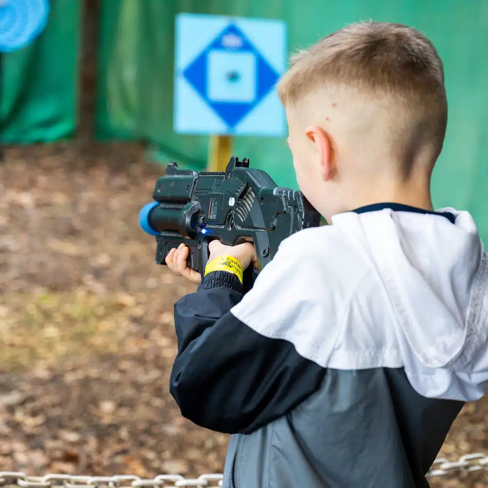 A young boy with short blond hair, wearing a grey and white jacket, aims a black toy laser gun at a blue and white target outdoors during the Navan Easter event. The ground is covered with leaves, with green barriers in the background.