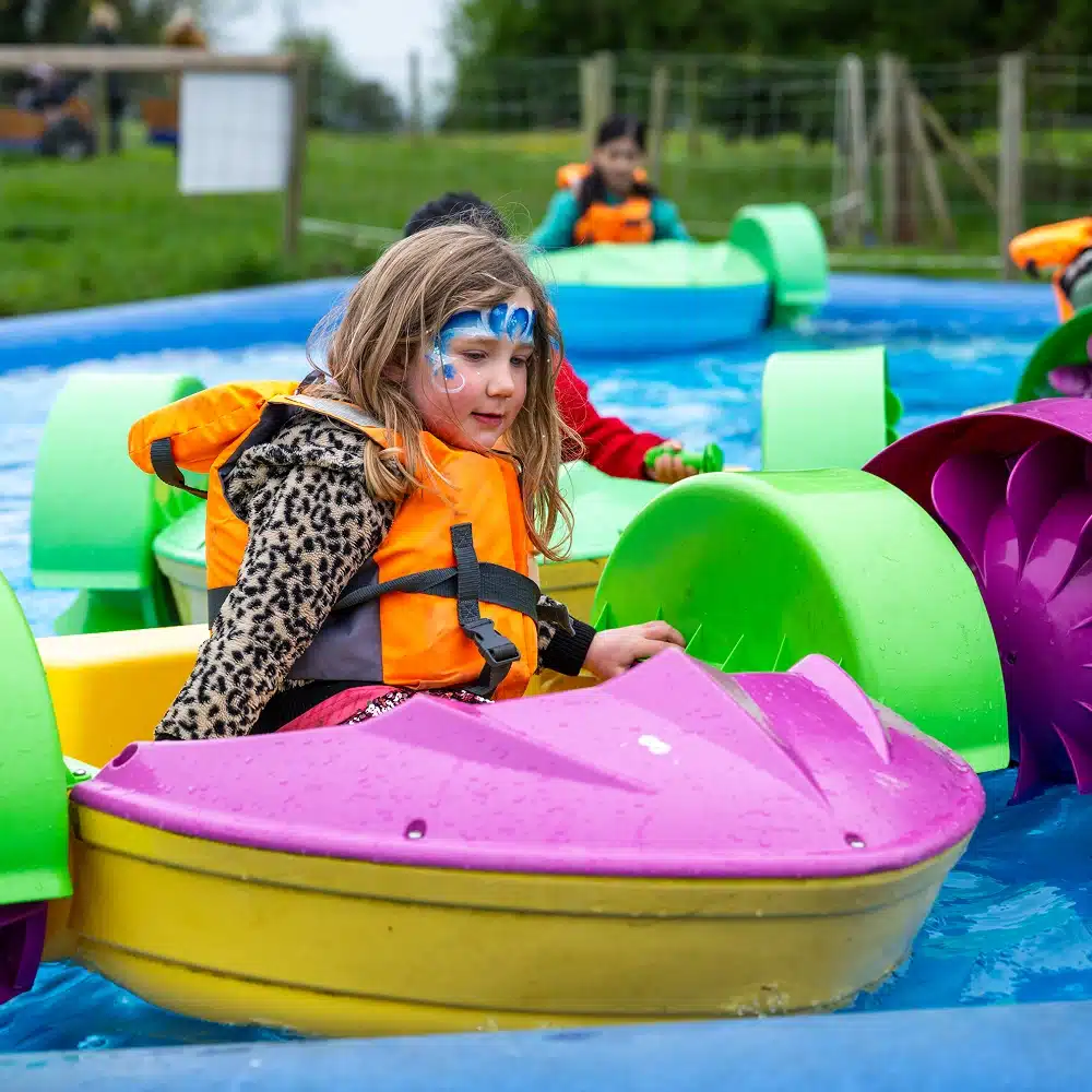 A young child with face paint and a life jacket rides a colourful paddle boat in a small pool at the Navan Easter event, with other children in similar boats enjoying the outdoor festivities on a cloudy day.