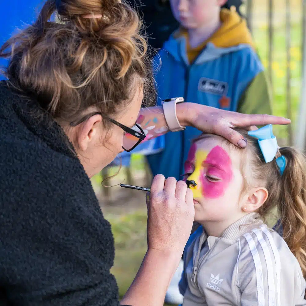 At the Navan Easter event, a woman paints a young girl’s face with bright pink and yellow colours. The girl has her eyes closed and a bow in her hair, whilst another child stands in the background watching.