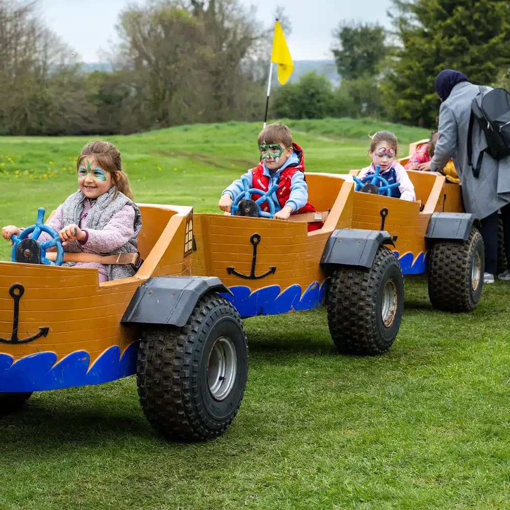Four young children with face paint are sitting in a row of small, yellow, boat-themed carts on grass, smiling and holding onto blue steering wheels. An adult stands nearby. Trees and a yellow flag are in the background.