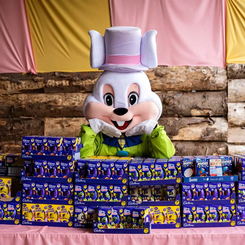 A person in a large white Easter Bunny costume with a top hat sits behind a display of stacked Cadbury Easter egg boxes, with pink and yellow fabric and a rustic wooden wall in the background.