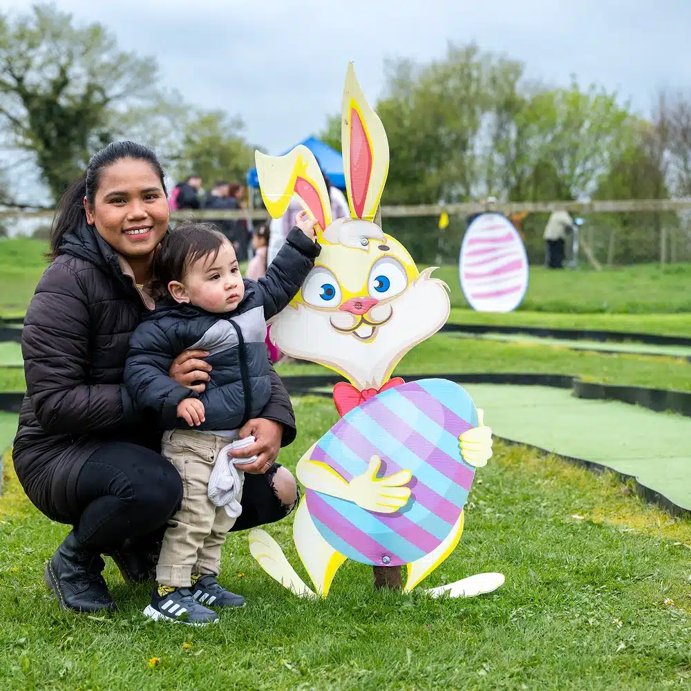 A woman and a young child pose outdoors next to a large cardboard Easter bunny holding a decorated egg. The child touches the bunny's ear, and both are smiling. Green grass and trees are visible in the background.