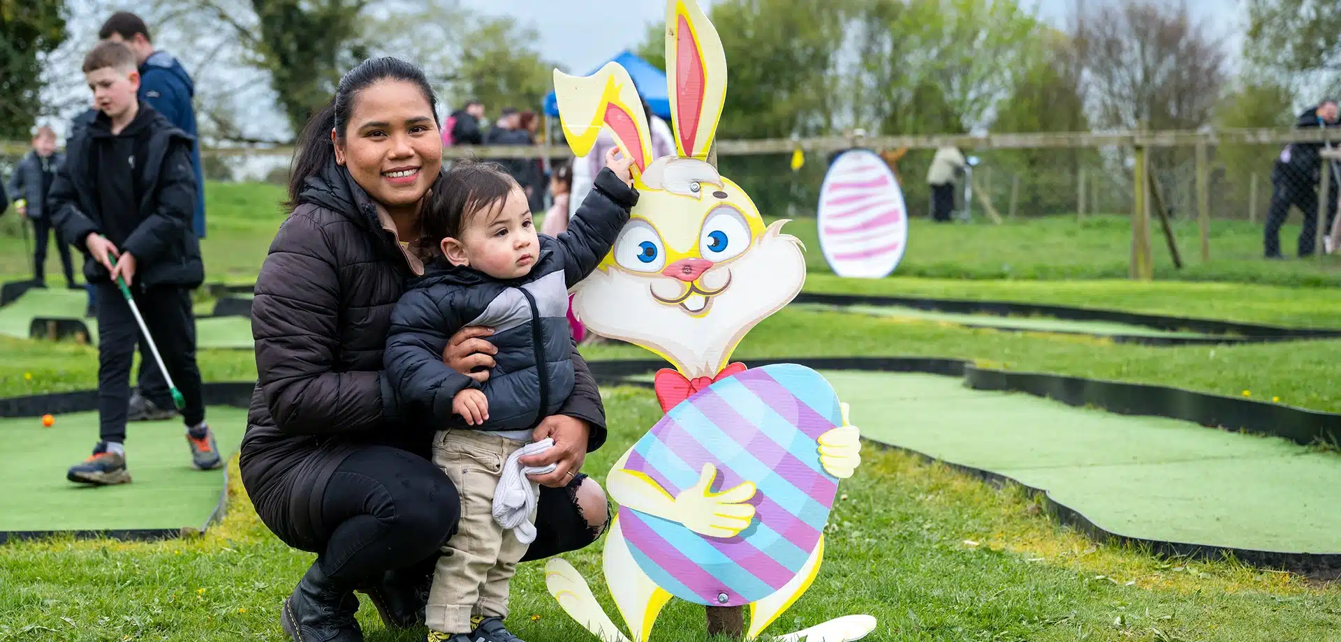 A woman and a young child pose next to a large cartoon bunny cut-out holding a decorated egg at an outdoor mini-golf course. Other people play golf in the background on a grassy field.