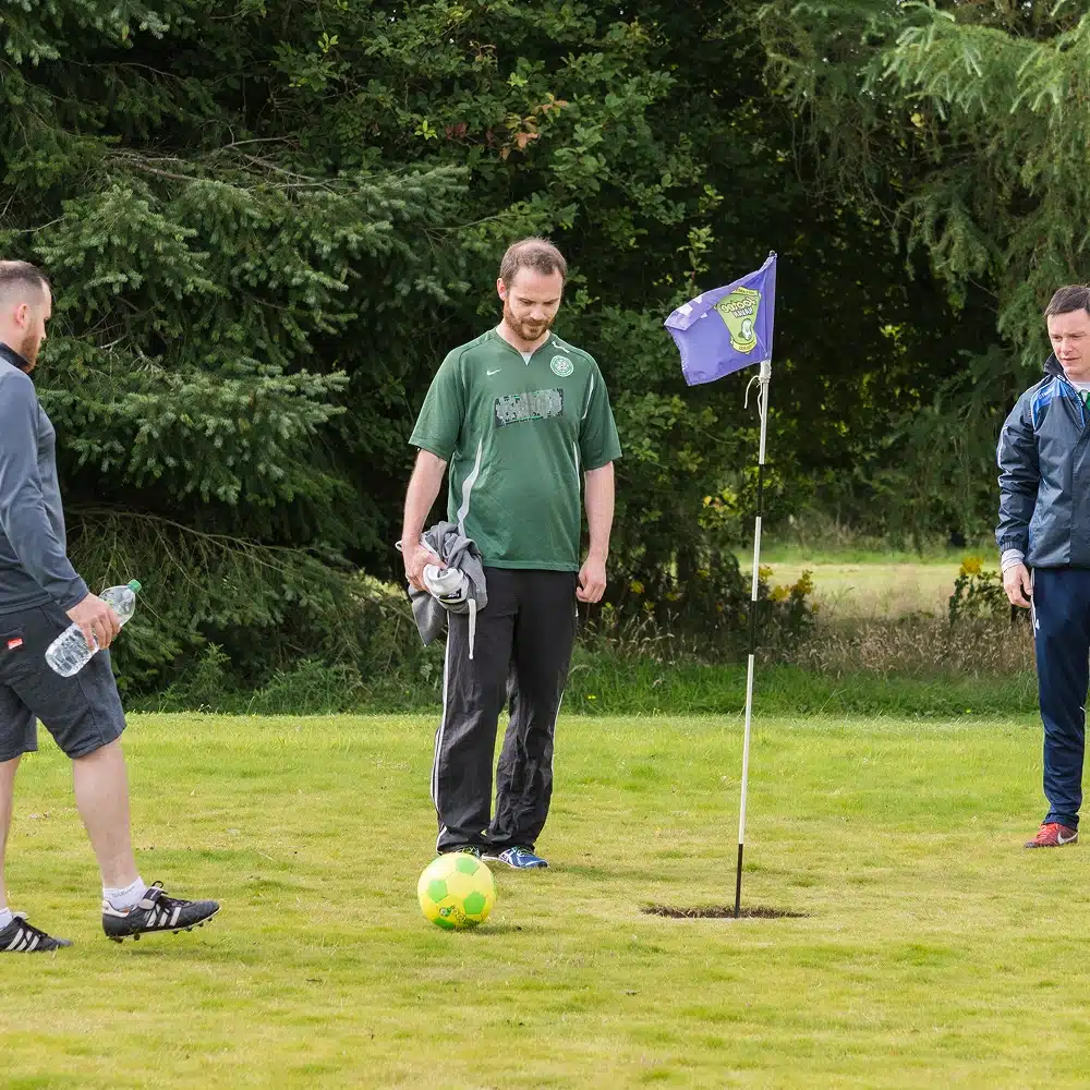 Three men stand on grassy ground near a yellow and green football and a flag marking a hole, suggesting they are playing Navan football golf. Trees are in the background, and the group looks relaxed outdoors.