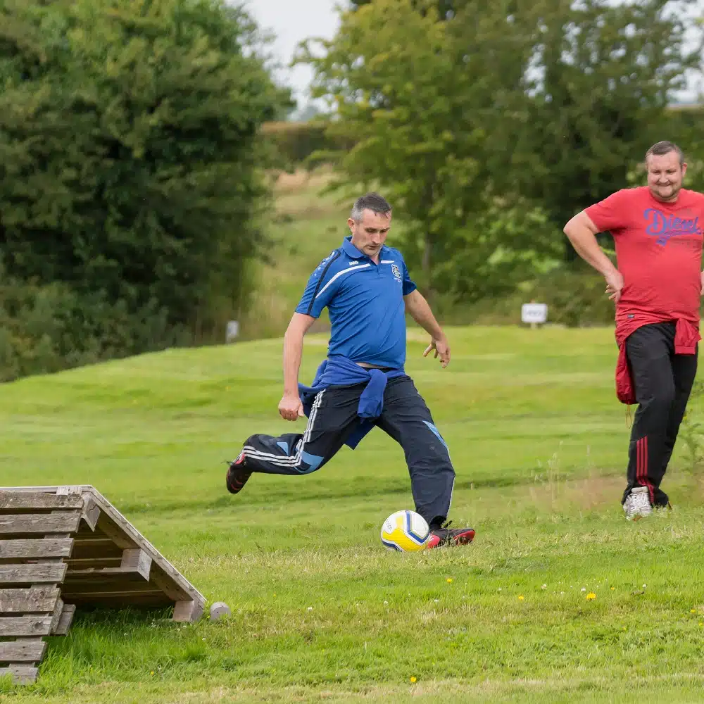 Two men are outdoors on a grassy field at Navan football golf. One man in a blue shirt is about to kick a football, while another in red stands nearby. A wooden ramp structure and trees are visible in the background.