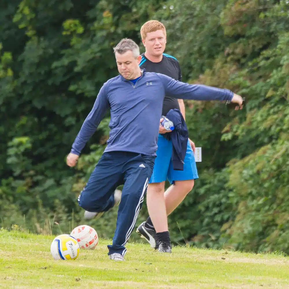 Two men are outdoors on grass; one in blue sportswear is kicking a Navan football golf ball, while the other stands behind him, watching and smiling. Trees fill the background.