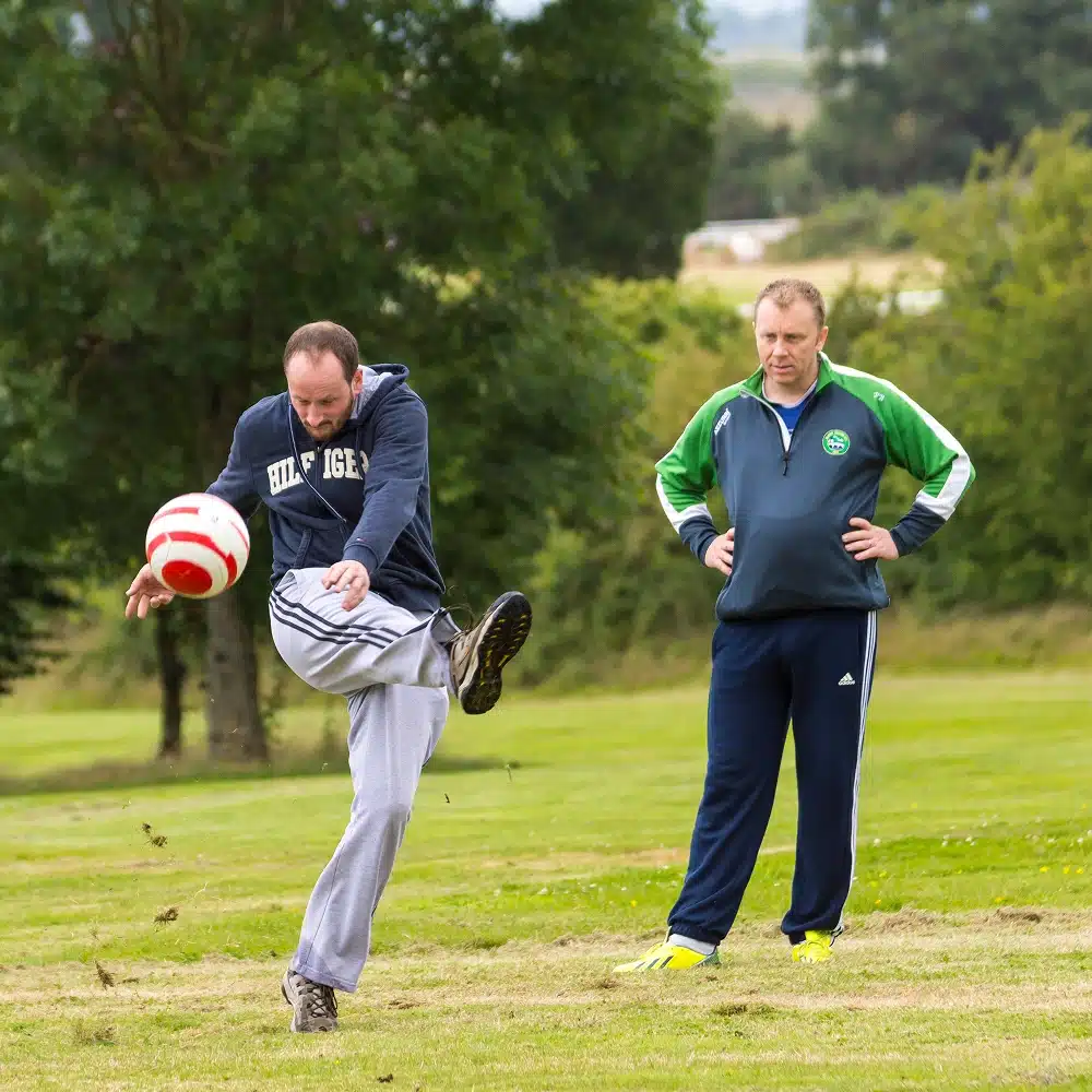 A man in a hoodie and tracksuit bottoms kicks a red and white ball on a grassy Navan football golf course, while another man in a tracksuit stands nearby watching. Trees and greenery are visible in the background.