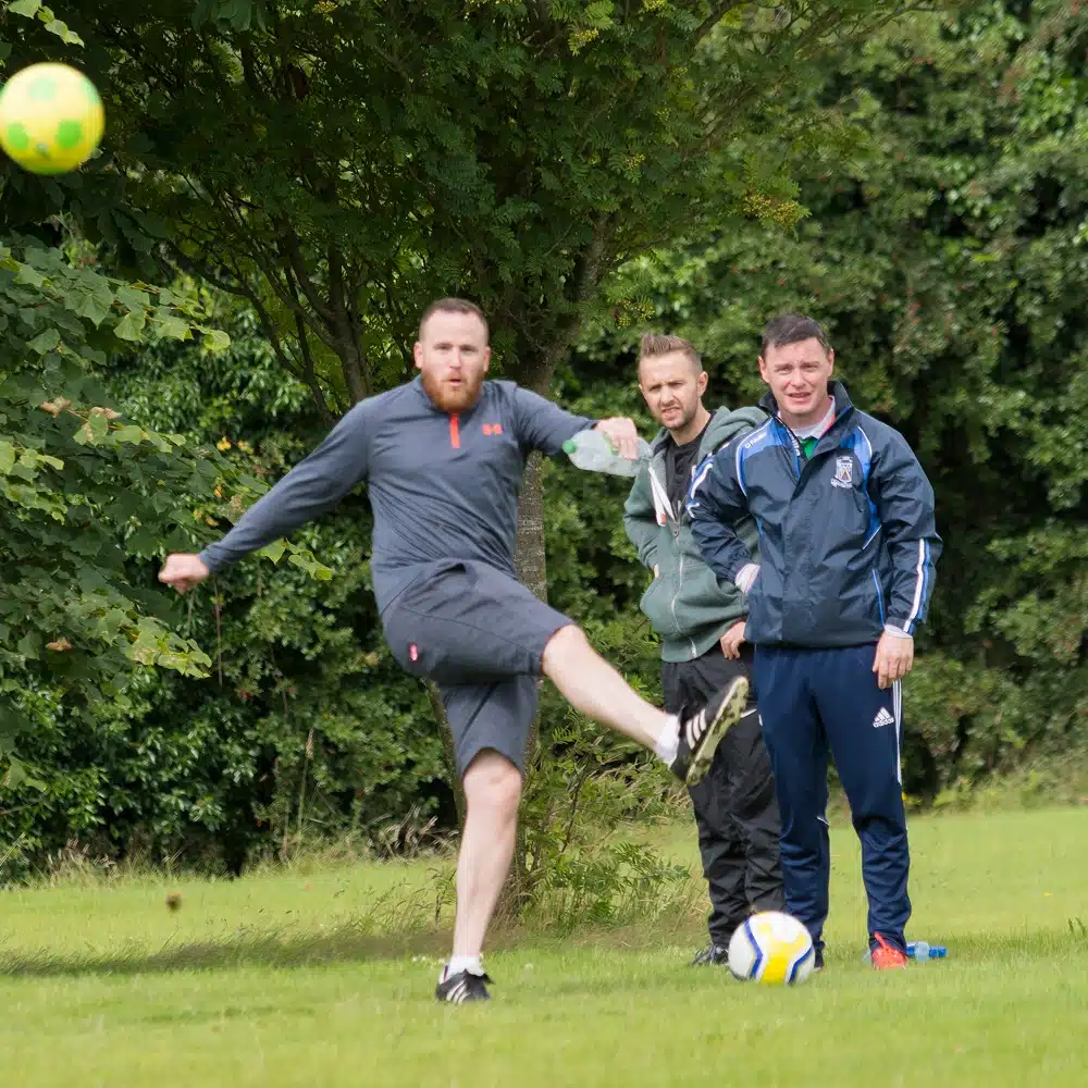 A man in sportswear kicks a football on a grassy field at Navan footgolf while two others watch in the background. Trees and greenery surround the scene, and another football lies at their feet.