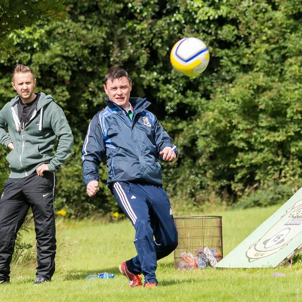 Two men stand on grass; one in a tracksuit kicks a football, while the other watches. With greenery and a tent behind them, the scene hints at a casual game of Navan football golf in an inviting outdoor setting.