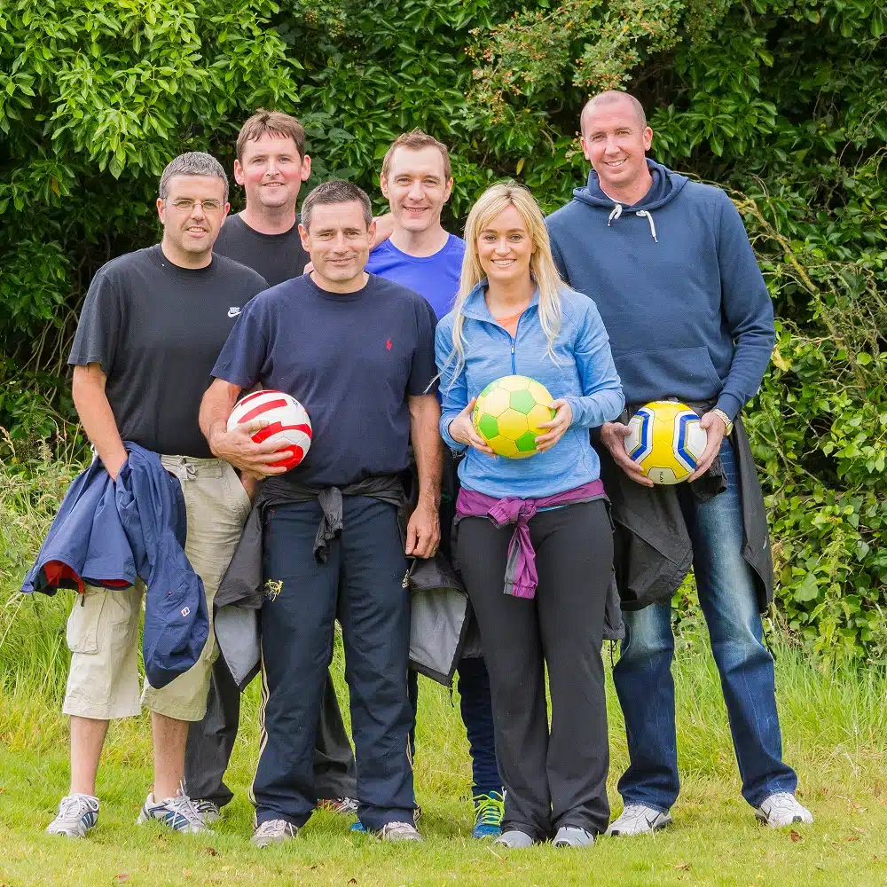 Six adults standing on grass outdoors, smiling at the camera. Three of them are holding differently coloured footballs from a Navan footgolf game. Green bushes are in the background, and everyone is dressed in casual clothing.