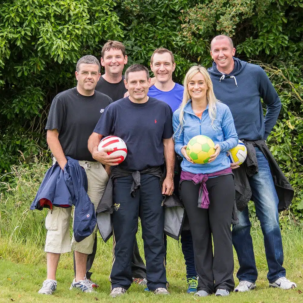 A group of six adults, four men and two women, stand together outdoors on grass at Navan football golf, smiling at the camera. Three are holding colourful footballs. Trees and bushes are in the background.