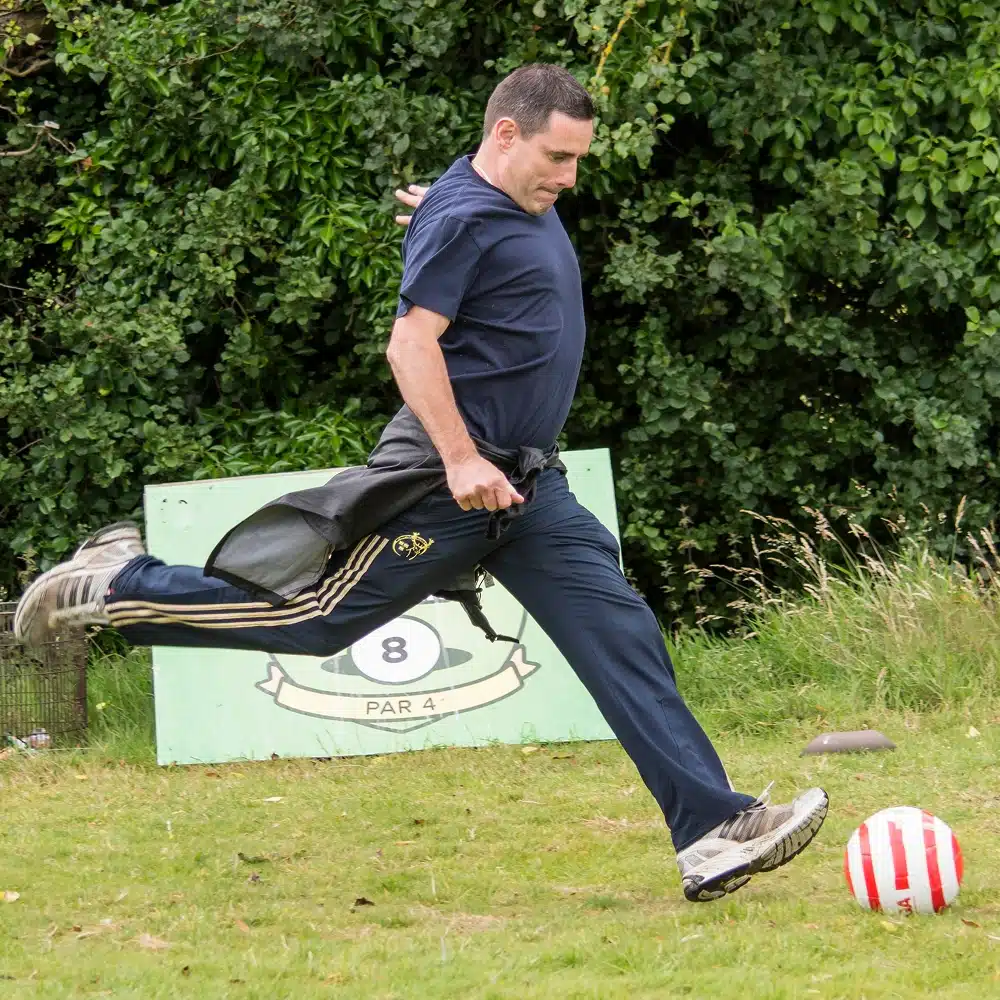 A man in a navy shirt and tracksuit bottoms kicks a red and white football outdoors at the Navan football golf course, with greenery and a sign labelled "8 Par 4" in the background.