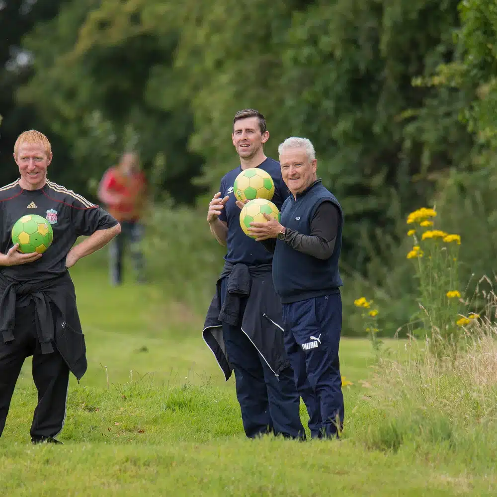 Three men standing on grass, each holding a green and yellow Navan football golf ball. They are smiling, dressed in sportswear, with trees and wildflowers visible in the background.