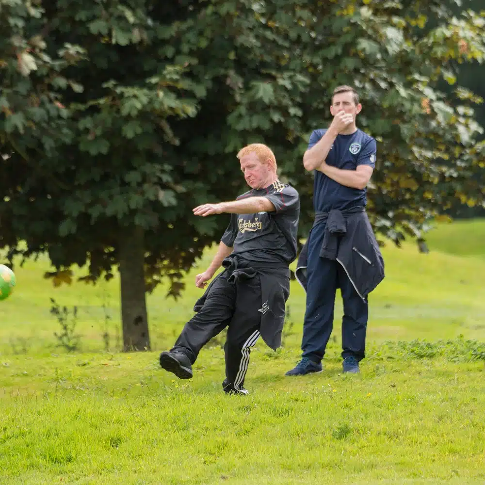 A man in black athletic clothing kicks a green ball on a grassy field during a game of Navan football golf, whilst another man in blue stands behind him watching with his hand on his chin. There are trees and greenery in the background.