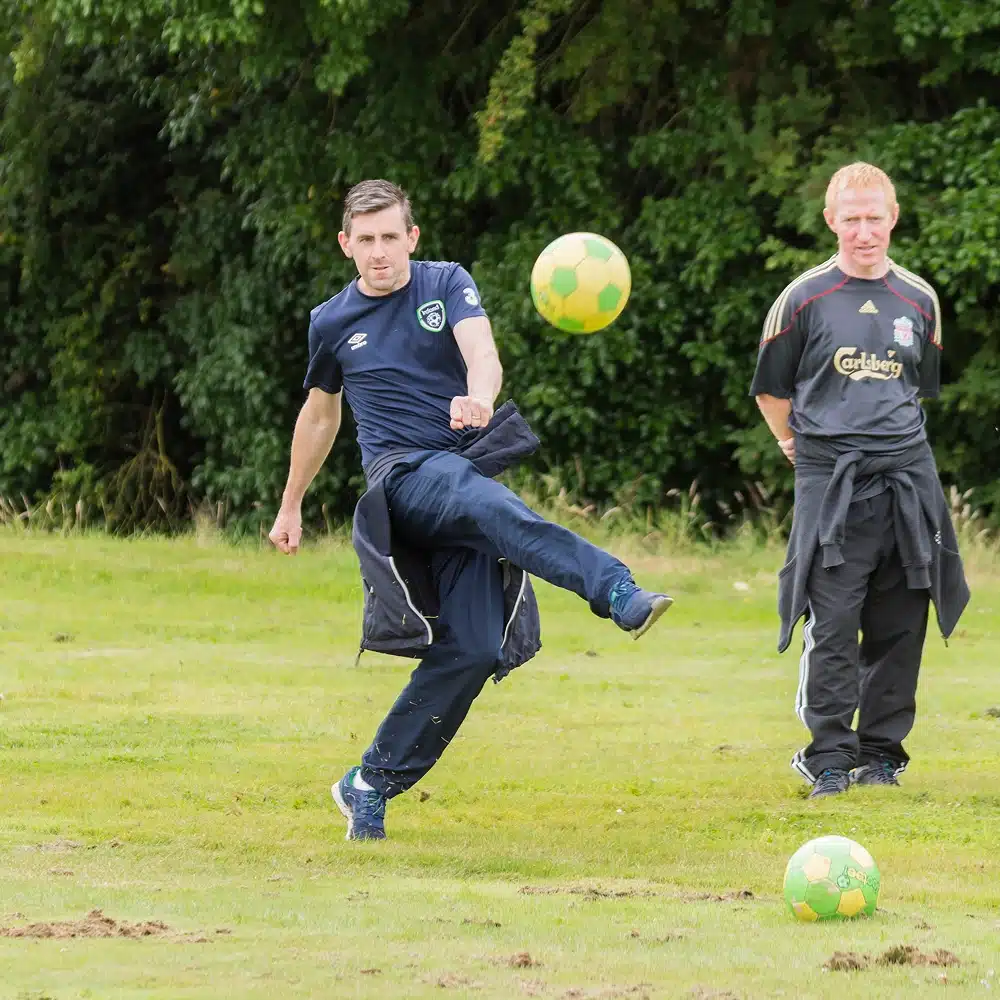 A man in a navy tracksuit kicks a yellow and green football on a grassy field, while another man watches in the background. Trees are visible behind them, evoking the casual spirit of Navan football golf.