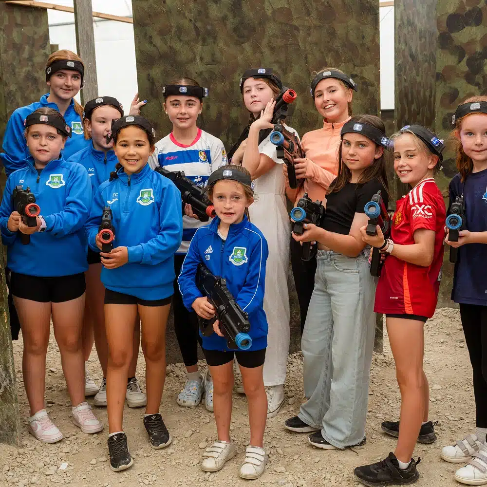A group of young girls from Navan secondary school, wearing sports clothing and headbands, pose together with laser tag guns in an indoor play area with camouflage barriers. They are smiling and appear to be enjoying their game.