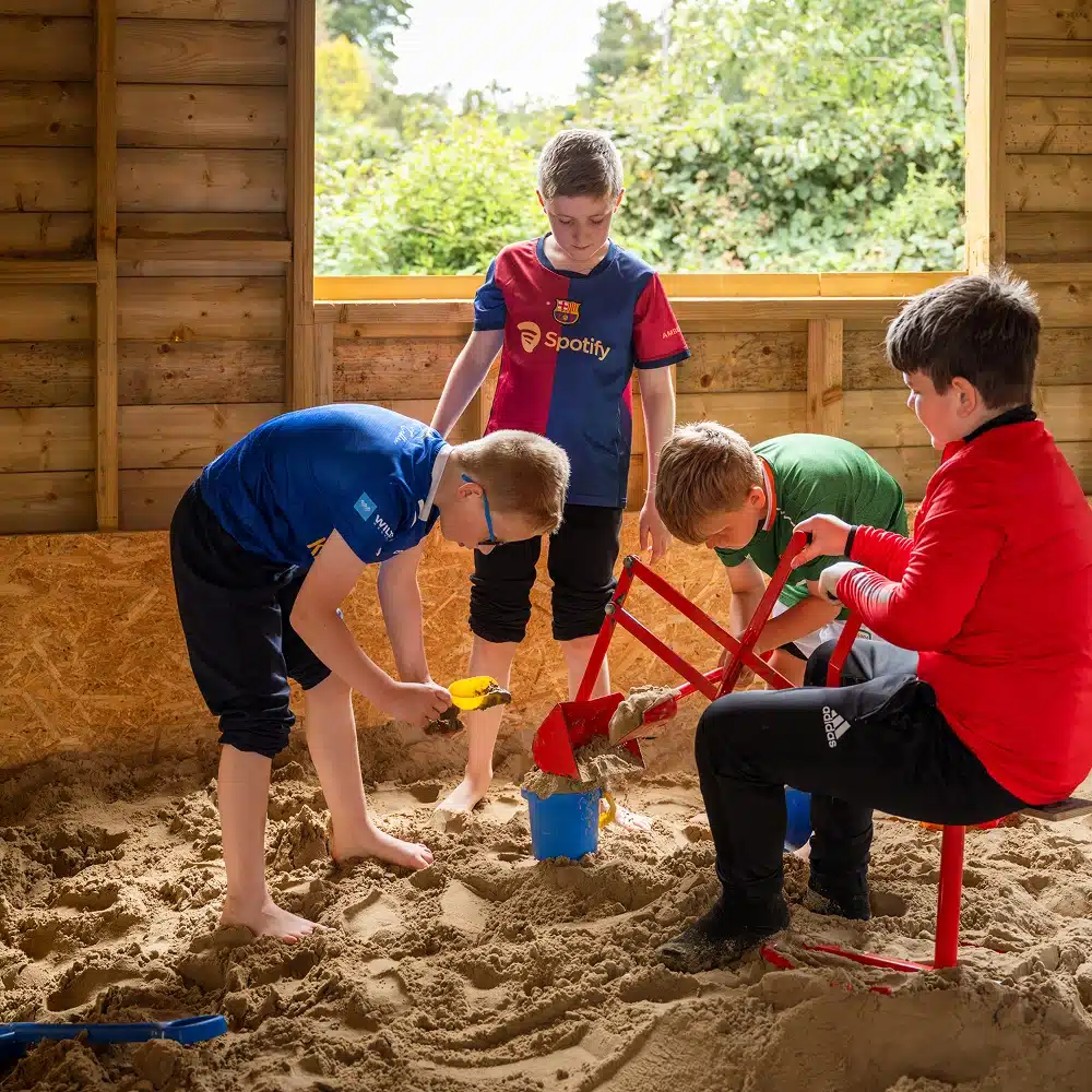 Four boys play together in an indoor sandpit at a Navan bouncy castle party, using spades and a digging tool to scoop sand into blue buckets. Sunlight streams through a large window, highlighting the wooden walls and green trees outside.