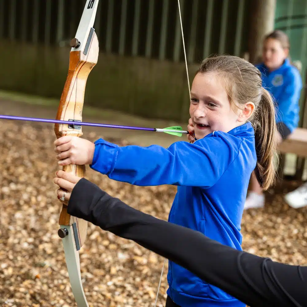 A young girl in a blue jacket smiles as she draws a bow and arrow, aiming at a target. She is outdoors on a woodchip surface, guided by an adult—her Navan secondary school group has just finished laser tag nearby.