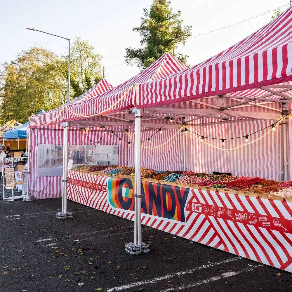 A colourful outdoor market stall with red-and-white striped tents displays a wide variety of sweets in clear bins. The stall is set up in a car park with trees in the background.