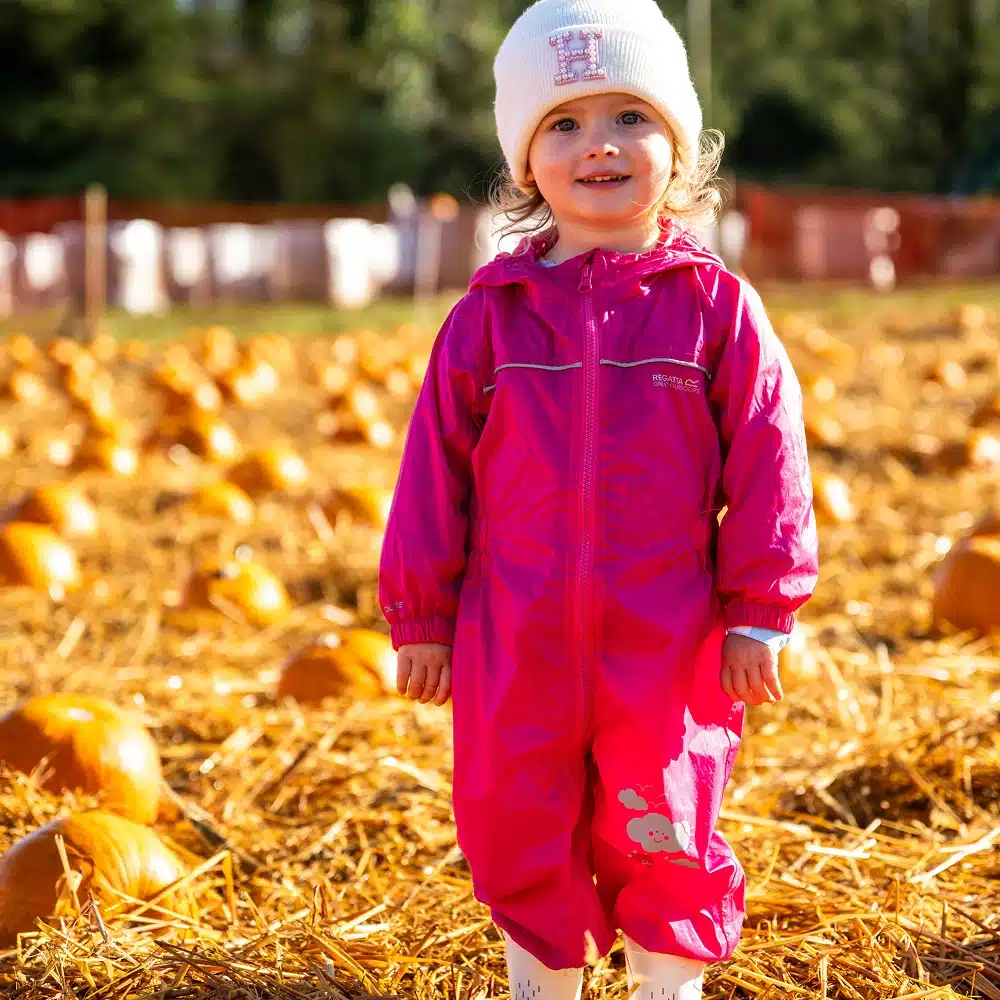 A young child in a bright pink rain suit and white beanie smiles whilst standing in a pumpkin patch filled with pumpkins and straw on a sunny day.