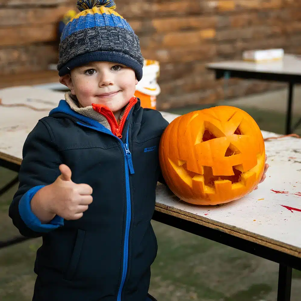 A young child in a blue winter jacket and hat gives a thumbs up whilst standing next to a carved pumpkin lantern on a table. The background is rustic with wooden walls and more tables.