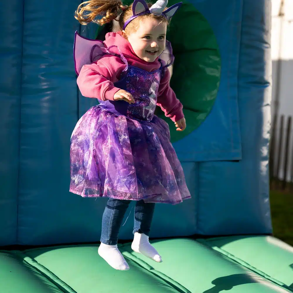 A young girl wearing a purple fairy dress, unicorn headband, and wings joyfully jumps on a green and blue inflatable bouncy castle outdoors, with sunlight shining on her.