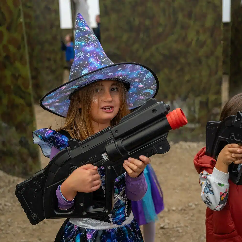 A young girl dressed as a witch, wearing a colourful pointed hat and holding a large toy blaster, stands outdoors in a play area with camouflage barriers. Another child holding a blaster is partly visible beside her.