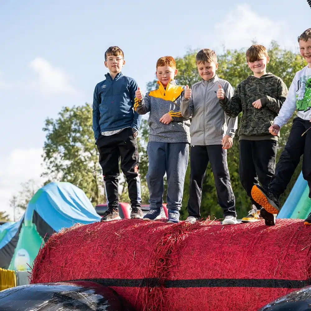 Five children stand on large red hay bales outdoors, smiling and giving thumbs up. Tents and trees are visible in the background, suggesting a fun event or camp. The sky is blue with scattered clouds.