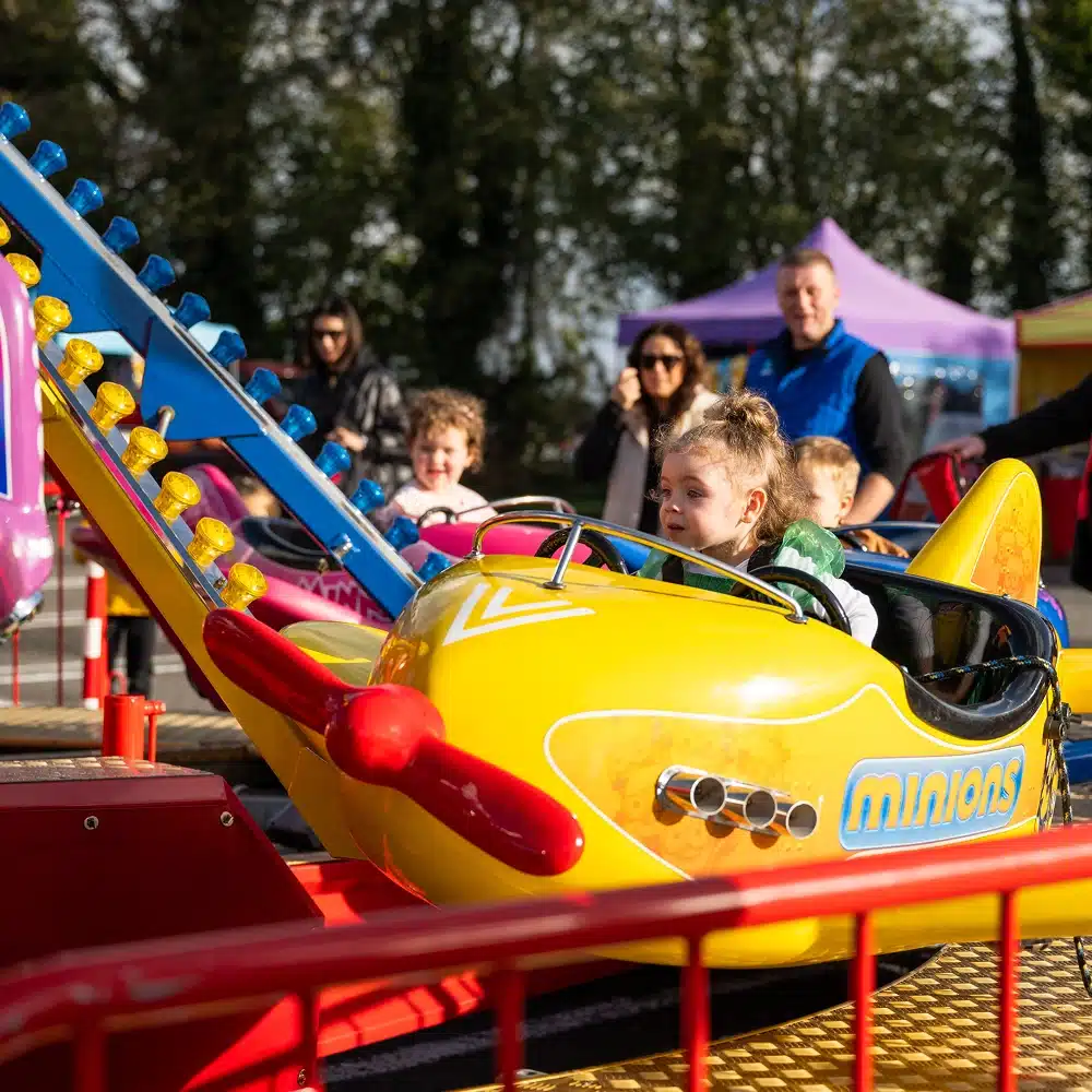 A young girl smiles whilst riding a yellow Minions-themed aeroplane ride at an outdoor fair, with other children and adults watching in the background. Bright sunlight and colourful carnival scenery are visible.