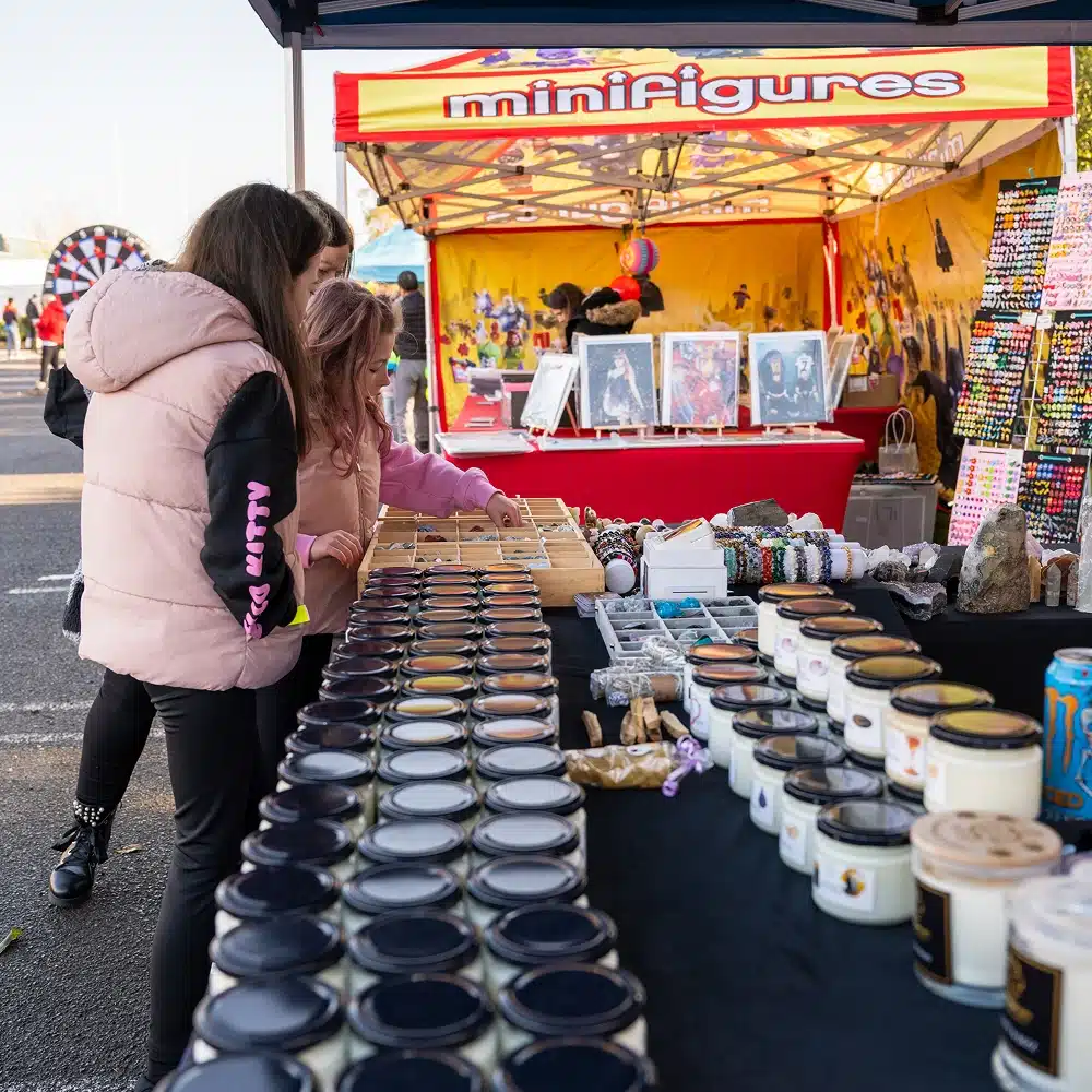 Two girls browse a market stall with candles, bracelets, and trinkets on display. The stall has a colourful tent with a sign reading “minifigures” and artwork in the background.