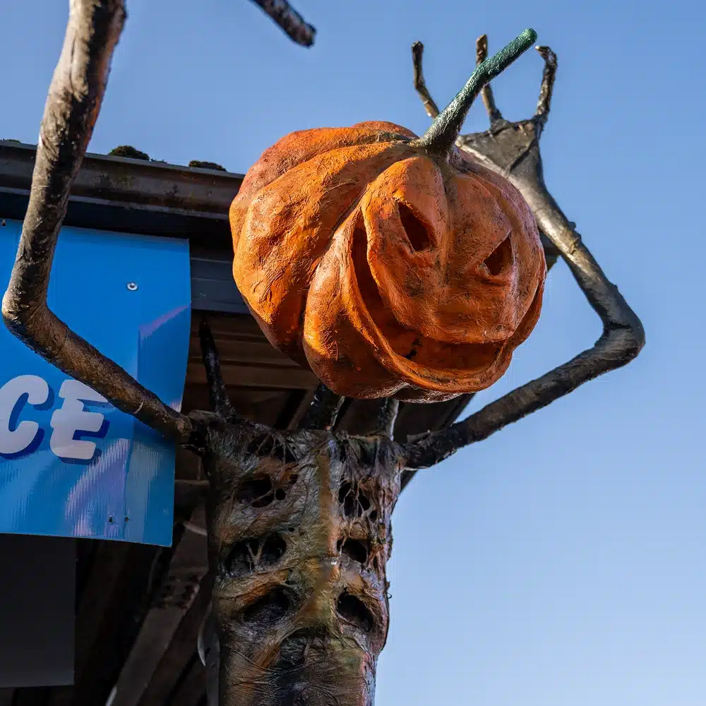 A large, creepy pumpkin creature with a carved, grinning face and long, twisted arms is mounted near a building against a clear blue sky.