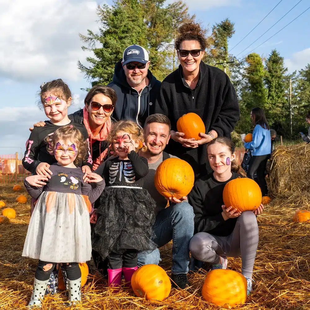 A group of smiling adults and children with painted faces pose together at a pumpkin patch, each holding a pumpkin. They are surrounded by hay and pumpkins, with trees and a blue sky in the background.