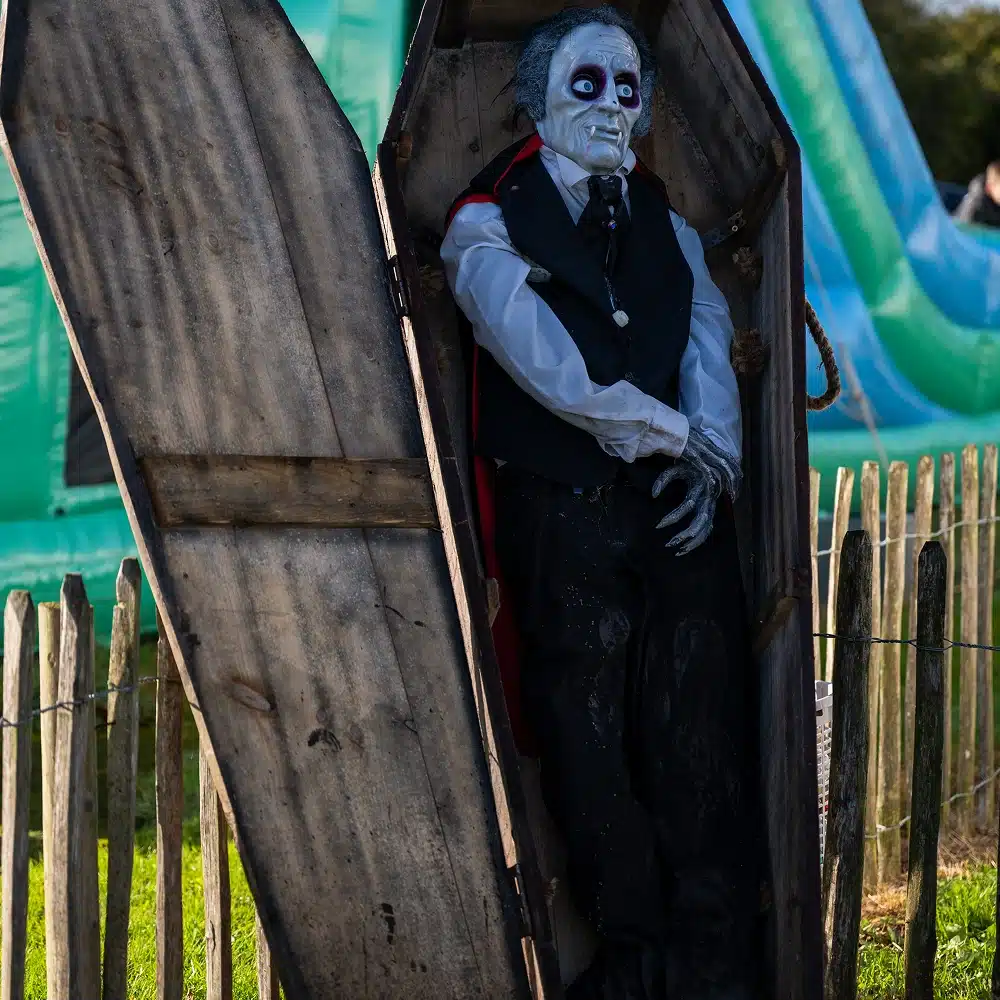A life-sized vampire mannequin with pale skin, dark eyes, and a formal suit stands in an upright wooden coffin outdoors, next to a picket fence and in front of an inflatable play structure.